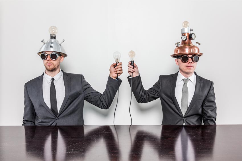 Imagen metafórica de lo que puede sentir un Interiorista Comercial: 2 personas con cascos con bombillas en sus manos y con cascos con bombillas y con medidores de aguja, que sostienen una bombilla en sus manos.